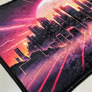 Puede incluir: Alfombra rectangular con un dise&ntilde;o de paisaje urbano retro. La obra de arte presenta un gran sol, rascacielos y l&iacute;neas radiantes en tonos rojos, morados y naranjas. La alfombra tiene un borde negro. El dise&ntilde;o evoca una ciudad futurista.