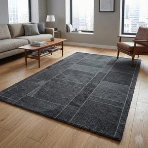 May include: A dark grey rectangular rug with a geometric pattern of squares and rectangles. The rug has a textured appearance and is bordered by a thin white line. The rug is placed on a wooden floor.