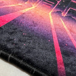 Puede incluir: Primer plano de una alfombra negra y afelpada con un dise&ntilde;o vibrante y colorido. El dise&ntilde;o presenta un paisaje urbano con tonos rosas, naranjas y morados, creando una est&eacute;tica retro y ne&oacute;n. La alfombra tiene un borde negro.