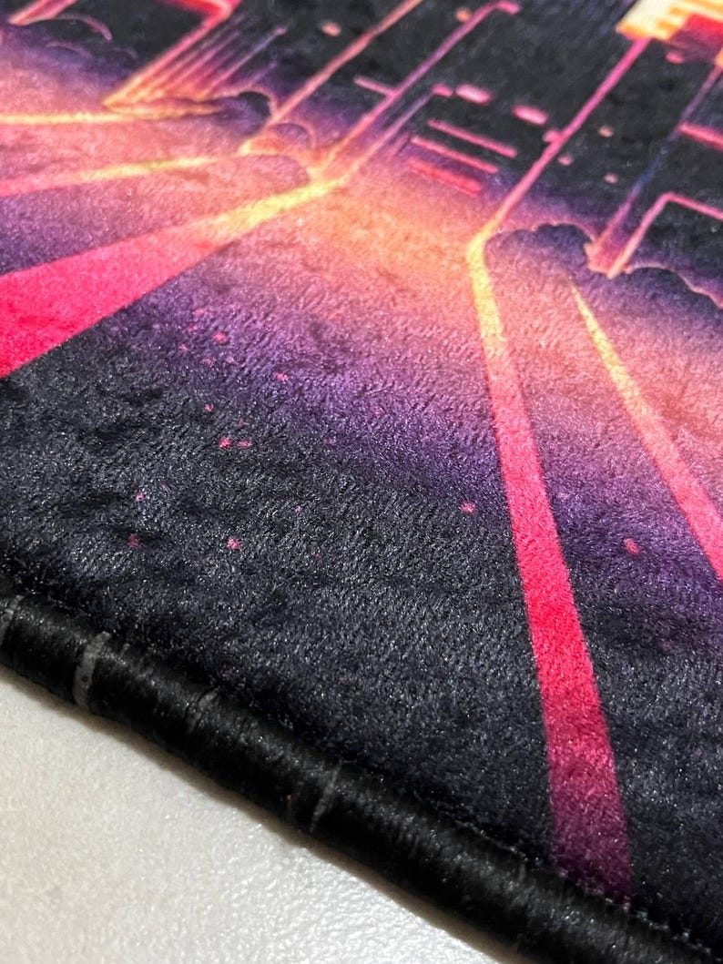 Puede incluir: Primer plano de una alfombra negra y afelpada con un dise&ntilde;o vibrante y colorido. El dise&ntilde;o presenta un paisaje urbano con tonos rosas, naranjas y morados, creando una est&eacute;tica retro y ne&oacute;n. La alfombra tiene un borde negro.