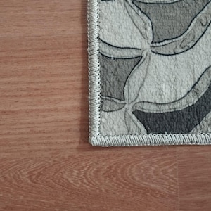 May include: A section of a patterned rug with a gray, black, and white design. The rug has a wavy, floral-like pattern and a light grey border. The rug is placed on a wooden floor.