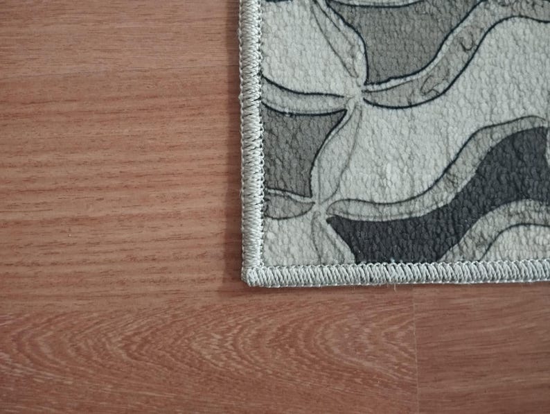May include: A section of a patterned rug with a gray, black, and white design. The rug has a wavy, floral-like pattern and a light grey border. The rug is placed on a wooden floor.