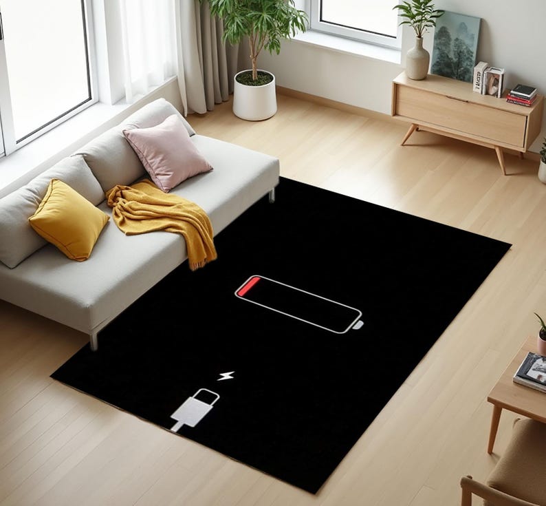 Puede incluir: Una alfombra rectangular negra con un contorno blanco de un s&iacute;mbolo de bater&iacute;a baja y una parte superior roja. Tambi&eacute;n hay un icono de cable de carga blanco. La alfombra est&aacute; sobre un suelo de madera clara, cerca de un sof&aacute; y una peque&ntilde;a mesa de madera.