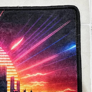 Puede incluir: Alfombrilla de rat&oacute;n rectangular con una silueta de ciudad contra un fondo de puesta de sol. El dise&ntilde;o incorpora tonos de naranja, rojo, morado y azul, con rayos de luz que emanan del sol. La alfombrilla tiene un borde negro.