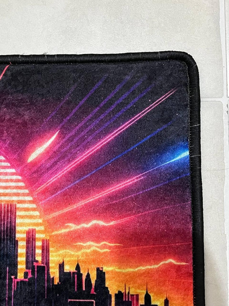 Puede incluir: Alfombrilla de rat&oacute;n rectangular con una silueta de ciudad contra un fondo de puesta de sol. El dise&ntilde;o incorpora tonos de naranja, rojo, morado y azul, con rayos de luz que emanan del sol. La alfombrilla tiene un borde negro.