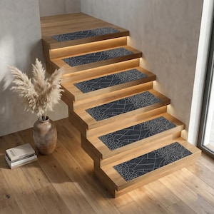 May include: Wooden staircase with built-in lighting and dark grey stair treads featuring a cracked pattern. A vase with dried plants and a stack of books are on the landing. The steps are made of light-coloured wood.