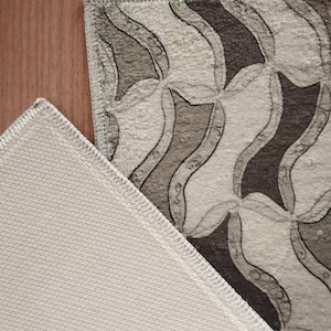 May include: A rectangular rug featuring a repeating pattern of curved shapes in shades of grey and white. The rug's edge is visible, showing a stitched border. The underside of the rug is a white, textured material.