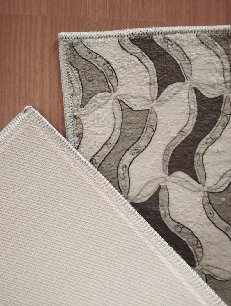 May include: A rectangular rug featuring a repeating pattern of curved shapes in shades of grey and white. The rug's edge is visible, showing a stitched border. The underside of the rug is a white, textured material.
