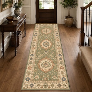 May include: A long rectangular runner rug featuring a sage green background with a central repeating medallion pattern in cream, terracotta, and navy blue. The rug has a wide cream border decorated with floral motifs and geometric accents.