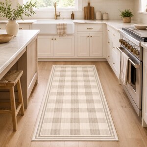 May include: A rectangular kitchen rug with a plaid pattern in shades of beige and gray. The rug has a border and is placed on a light-colored wooden floor. The kitchen features white cabinets, a farmhouse sink, and a stainless steel stove.