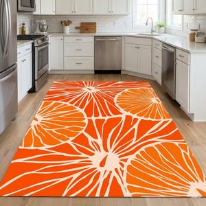 Puede incluir: Una alfombra rectangular con un vibrante estampado floral naranja y blanco. La alfombra está colocada sobre un suelo de madera en una cocina. El diseño presenta grandes rodajas de naranja estilizadas.