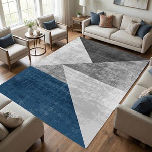 May include: A large area rug with a geometric design. The rug features a pattern of triangles in shades of blue, gray, and white. The rug is placed on a hardwood floor in a living room setting, with furniture visible in the background.