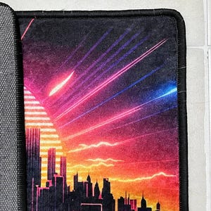 Puede incluir: Alfombrilla de rat&oacute;n rectangular con una silueta de ciudad contra un fondo de puesta de sol. El dise&ntilde;o presenta un degradado de colores naranja, rosa y morado, con rayos de luz. La alfombrilla tiene un borde negro.