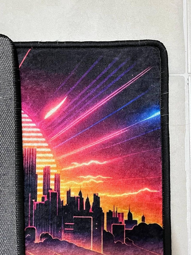 Puede incluir: Alfombrilla de rat&oacute;n rectangular con una silueta de ciudad contra un fondo de puesta de sol. El dise&ntilde;o presenta un degradado de colores naranja, rosa y morado, con rayos de luz. La alfombrilla tiene un borde negro.