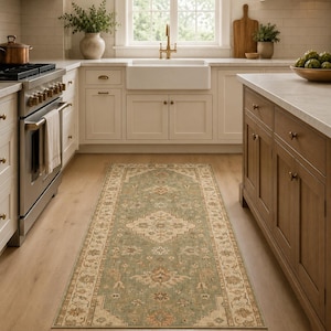 May include: A rectangular rug with a distressed appearance, featuring a central floral medallion. The rug has a sage green field with beige and rust accents. The border displays a repeating floral pattern. The rug is in a kitchen with light wood flooring.
