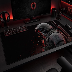 Può includere: Tappetino per mouse da gioco nero con un design a soldato con cappuccio e teschio. Il tappetino ha una superficie nera con un'illustrazione rossa e grigia. Sono visibili anche una tastiera, delle cuffie e un monitor.