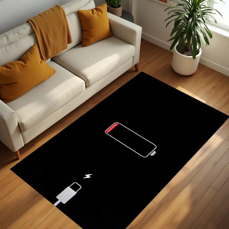 Puede incluir: Alfombra rectangular negra con un contorno blanco de un s&iacute;mbolo de bater&iacute;a baja con la parte superior roja y un cable de carga blanco. La alfombra est&aacute; sobre un suelo de madera, cerca de un sof&aacute; blanco con cojines amarillos y una planta de interior.