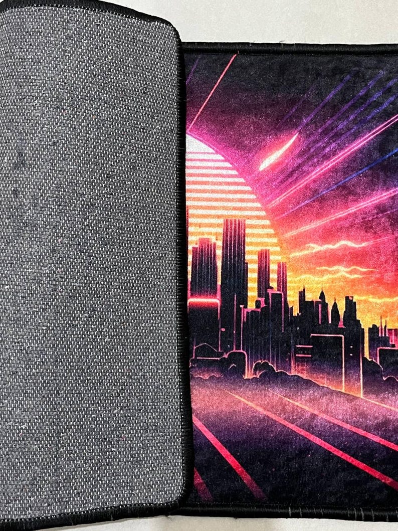 Puede incluir: Alfombrilla de rat&oacute;n negra con un dise&ntilde;o retro de paisaje urbano. La imagen presenta una vibrante puesta de sol con tonos rosas y naranjas, una silueta del horizonte de la ciudad y rayos de luz. La parte posterior de la alfombrilla es gris.