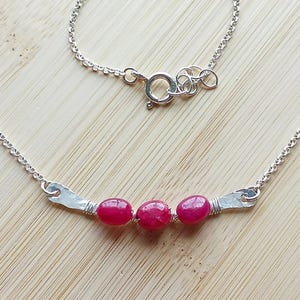 May include: A silver necklace featuring three oval, bright red gemstones. The gemstones are set in a hammered silver bar, with a delicate chain and clasp. The necklace is displayed on a light wood surface.