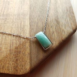 May include: A silver chain necklace featuring a rectangular pendant with a smooth, light green gemstone. The pendant is held within a thin silver frame that connects directly to the delicate metal chain links on both sides of the stone.