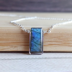 May include: A rectangular pendant necklace featuring a blue and green iridescent gemstone set in a silver-colored metal frame. The pendant hangs from a delicate silver chain, creating a simple yet elegant design. The gemstone's color shifts with light.