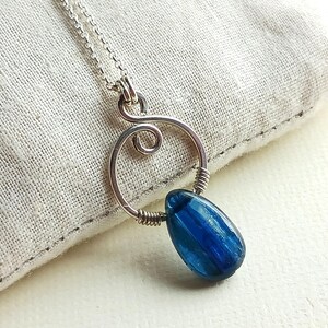 May include: A silver-toned necklace with a pendant featuring a vibrant blue, teardrop-shaped gemstone. The pendant is suspended from a delicate chain and has a unique, wire-wrapped design.