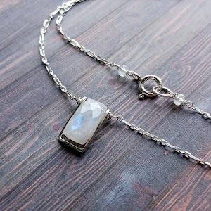 May include: A silver necklace featuring a rectangular, faceted moonstone pendant. The pendant is set in a silver frame and hangs from a delicate chain. The moonstone displays a subtle play of blue and white colors.