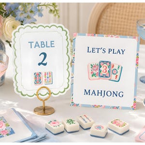 May include: A scalloped table number card labeled TABLE 2 and a rectangular sign reading LET'S PLAY MAHJONG, both featuring floral illustrations. Five white mahjong tiles with floral and character designs are arranged on the table surface.