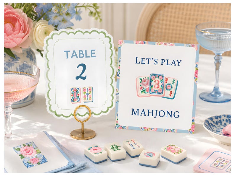 May include: A scalloped table number card labeled TABLE 2 and a rectangular sign reading LET'S PLAY MAHJONG, both featuring floral illustrations. Five white mahjong tiles with floral and character designs are arranged on the table surface.