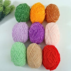 May include: Nine skeins of yarn in various colors, including green, orange, brown, lavender, purple, pink, light green, tan, and red. The yarn is arranged in three rows of three, with each skein appearing to be made of a soft, textured material.