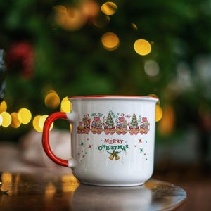 May include: White enamel mug with a red rim and handle. The mug features a festive design of Christmas trees and trains, with the words "MERRY CHRISTMAS" printed on the front. The background is blurred with Christmas lights.