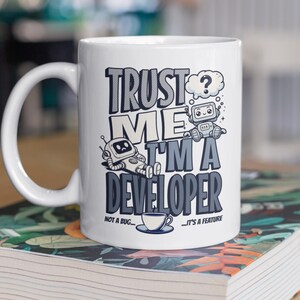 Op de afbeelding: Witte keramische mok met de tekst "TRUST ME I'M A DEVELOPER" in een zwart, grijs en wit ontwerp. De mok heeft cartoonrobots en een koffiekopje. De zin "NOT A BUG... IT'S A FEATURE" staat ook op de mok.