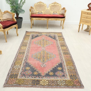 Puede incluir: Una alfombra rectangular de estilo vintage con un campo central rosa y patrones geométricos intrincados en azul marino, dorado y amarillo. La alfombra está colocada sobre un suelo de madera clara, con muebles antiguos en el fondo.