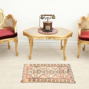 May include: Two gold-painted chairs with cane backrests and deep red velvet cushions, a matching gold-painted coffee table with a glass top, a vintage rotary telephone, and a small rectangular rug with a geometric pattern in pink and black.