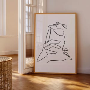 Op de afbeelding: Een ingelijste kunstdruk met een minimalistische zwarte lijntekening van een gezicht en een hand op een witte achtergrond. Het kunstwerk is ingelijst in een lichtbruine houten lijst, wat een moderne uitstraling creëert. De print wordt tentoongesteld in een goed verlichte kamer met houten vloeren.