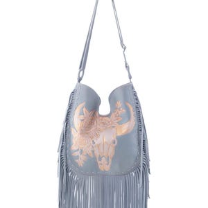 May include: A light blue shoulder bag with a decorative bull skull and floral design in gold. The bag has long fringe along the bottom and a long shoulder strap. The bag is made of a soft material.