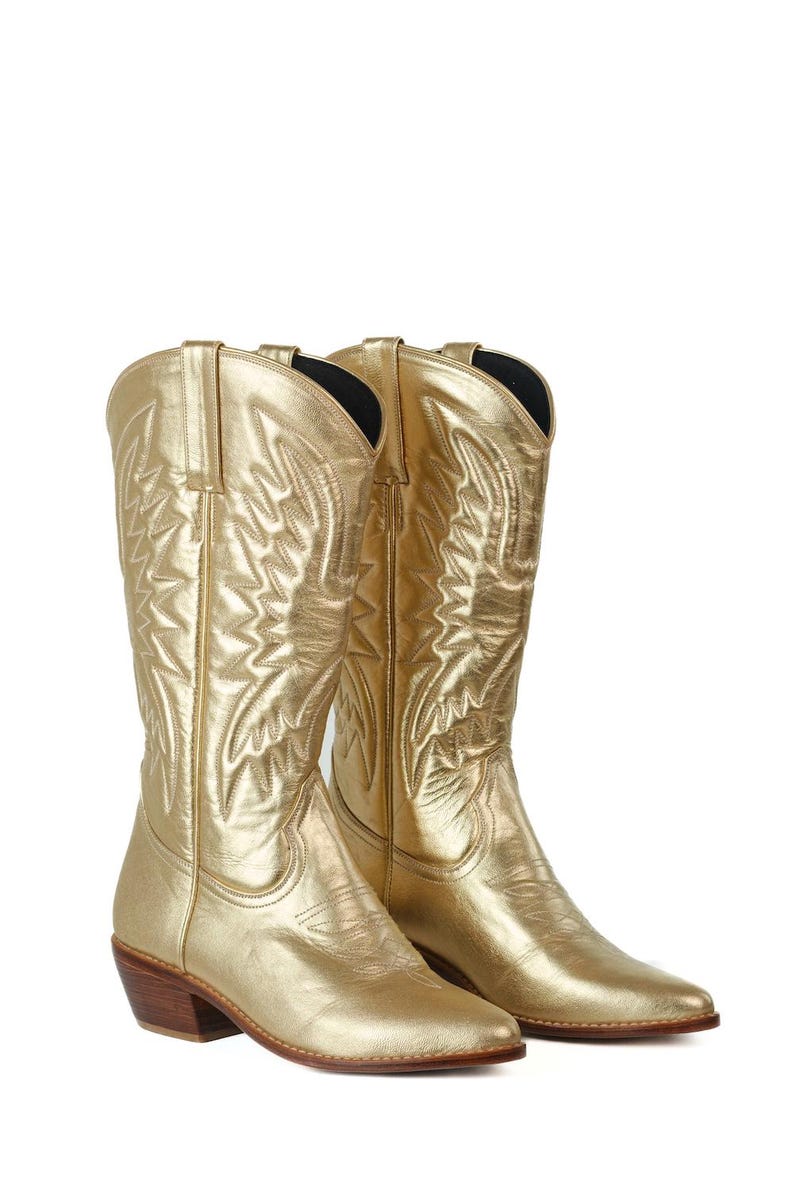Puede incluir: Un par de botas vaqueras de color dorado met&aacute;lico con punta afilada, tac&oacute;n de madera apilado y patrones de costura tonales intrincados en la ca&ntilde;a y el pie. Las botas tienen tiradores en la parte superior y un acabado liso y reflectante.