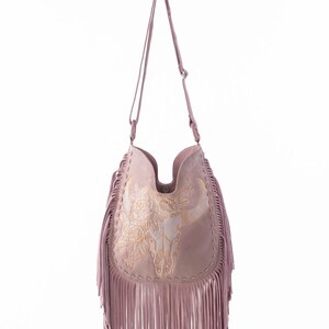 May include: Dusty rose-colored shoulder bag with a decorative bull skull and floral embroidery. The bag features long fringe along the bottom and a long shoulder strap. The bag is made of a soft, suede-like material.