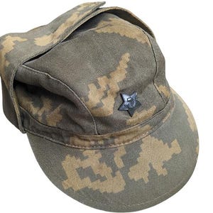 Puede incluir: Gorra militar con estampado de camuflaje en verde oliva y marrón. Presenta un pequeño emblema oscuro en forma de estrella. La visera es curva y la gorra tiene un diseño estructurado.