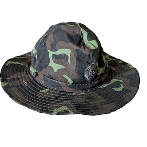 Op de afbeelding: Een hoed met een brede rand en een camouflagepatroon in de kleuren donkergroen, bruin en zwart. De hoed is voorzien van een rond metalen embleem aan de voorzijde, een stoffen band rond de kroon en kleine ventilatiegaatjes aan de zijkanten.