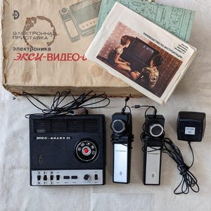 Op de afbeelding: Vintage zwarte en zilveren videogameconsole met twee controllers, een voedingsadapter en een doos. De doos heeft Russische tekst en een diagram van de console. De controllers zijn rechthoekig met een ronde knop. De console heeft een rode knop en diverse kleinere knoppen.