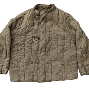 Peut inclure: Une veste matelassée beige avec un col montant, des manches longues et une fermeture boutonnée verticale. Le vêtement présente une surface texturée et froissée avec des coutures verticales, deux poches avant et des boutons aux poignets.