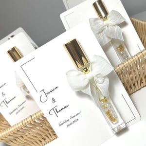 May include: Wedding favours comprising small perfume bottles with gold caps, decorated with white bows and filled with pearls and gold accents. Each bottle is attached to a white card with the names "Jessica & Thomas" and the wedding date.