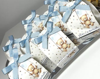 May include: Gift tags featuring a teddy bear on a swing design, tied with light blue ribbons. The tags are decorated with small grey polka dots and have the text "Nick's 1st Birthday" printed on them. A charming set for a first birthday.