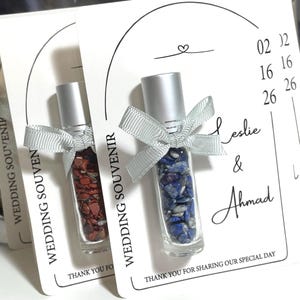May include: Two glass bottles filled with red and blue stones, with silver caps and ribbons. The bottles are on cards that say "WEDDING SOUVENIR" and "THANK YOU FOR SHARING OUR SPECIAL DAY". The cards also include the names "Leslie & Ahmad".