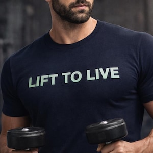 May include: Navy blue t-shirt with the text "LIFT TO LIVE" in a light green color. The person in the image is holding two black dumbbells. The background is blurred, suggesting a gym setting. The t-shirt is short-sleeved.