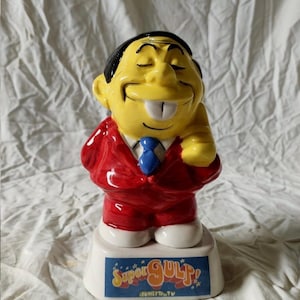 May include: A ceramic figurine of a yellow-skinned character with black hair, wearing a red suit and blue tie. The character has a wide grin with visible teeth. The base is white and features a blue rectangular label with the text Super GULP!.