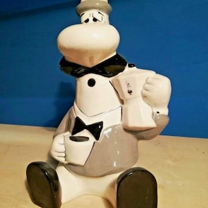 Op de afbeelding: Een keramieken beeldje van een stripfiguur met een grijze hoed, een zwarte vlinderdas en een grijs jasje. Het figuur heeft een wit gezicht, zwarte schoenen en houdt een witte koffiekop in de ene hand en een witte mokkapot in de andere.