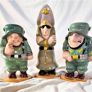 May include: Three ceramic figurines depicting cartoon soldiers. The figures are painted in shades of green, tan, and brown, with black accents. Each figure has a unique facial expression and is wearing a military uniform. The figurines are standing on small, round bases.