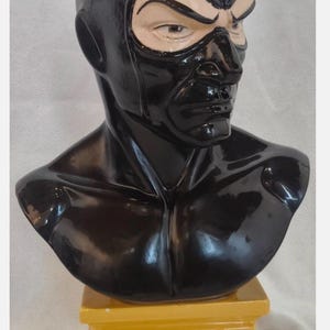 Op de afbeelding: Een glanzend zwart keramieken borstbeeld van een personage met een nauwsluitend masker dat het hoofd en de nek bedekt, waarbij de ogen en de huid eromheen zichtbaar zijn. Het beeld staat op een gele rechthoekige voet met het woord DIABOLIK in rode letters.